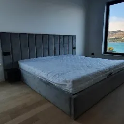 Szare tapicerowane łóżko z wysokim wezgłowiem z pionowymi panelami, ustawione w sypialni z widokiem na morze z dwóch okien.