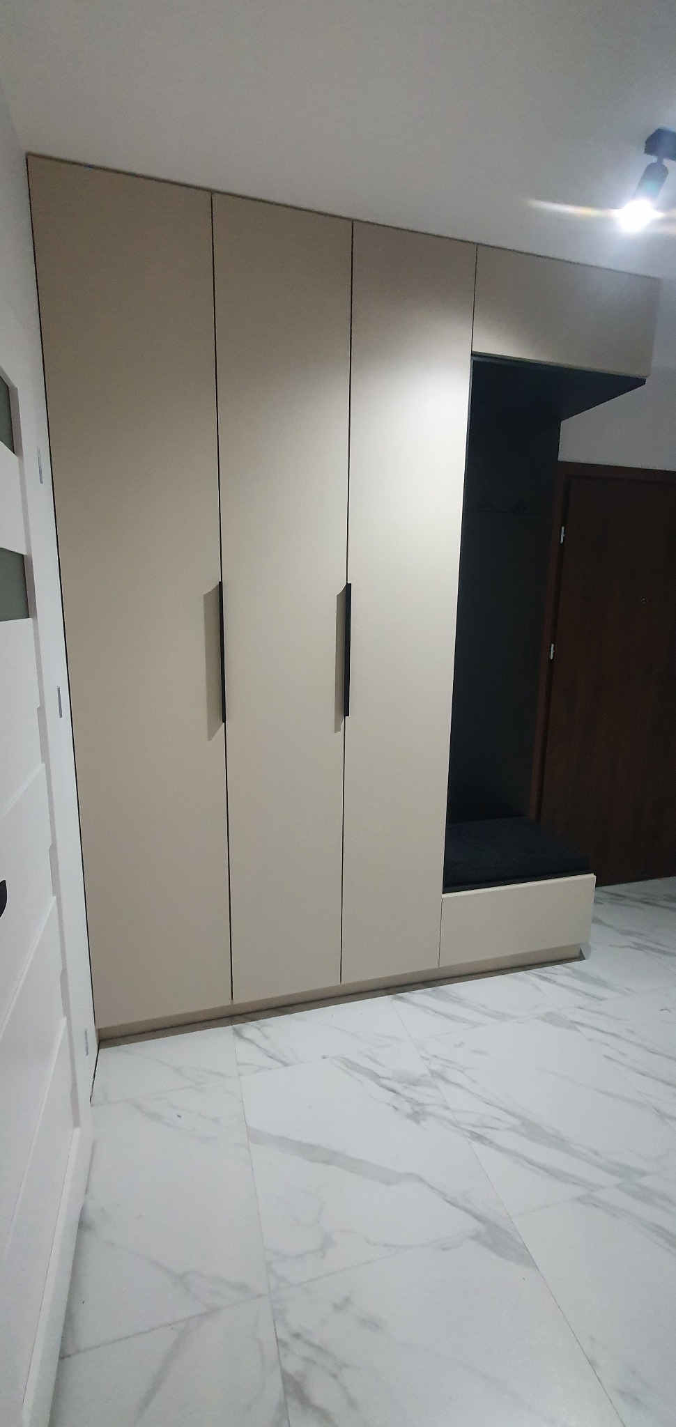 Beżowa szafa z trzema drzwiami i wbudowanym siedziskiem z czarną poduszką, uzupełniająca aranżację przedpokoju z marmurową podłogą.