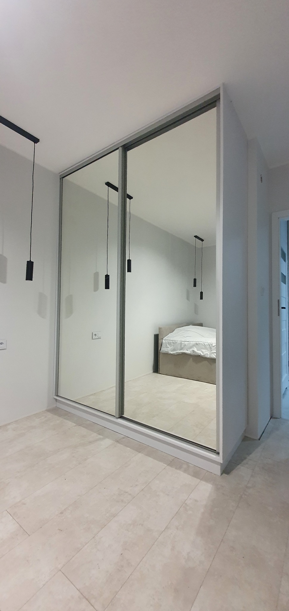 Nowoczesna szafa wnękowa z przesuwnymi lustrzanymi drzwiami, odzwierciedlająca wnętrze sypialni z minimalistycznym oświetleniem i łóżkiem.