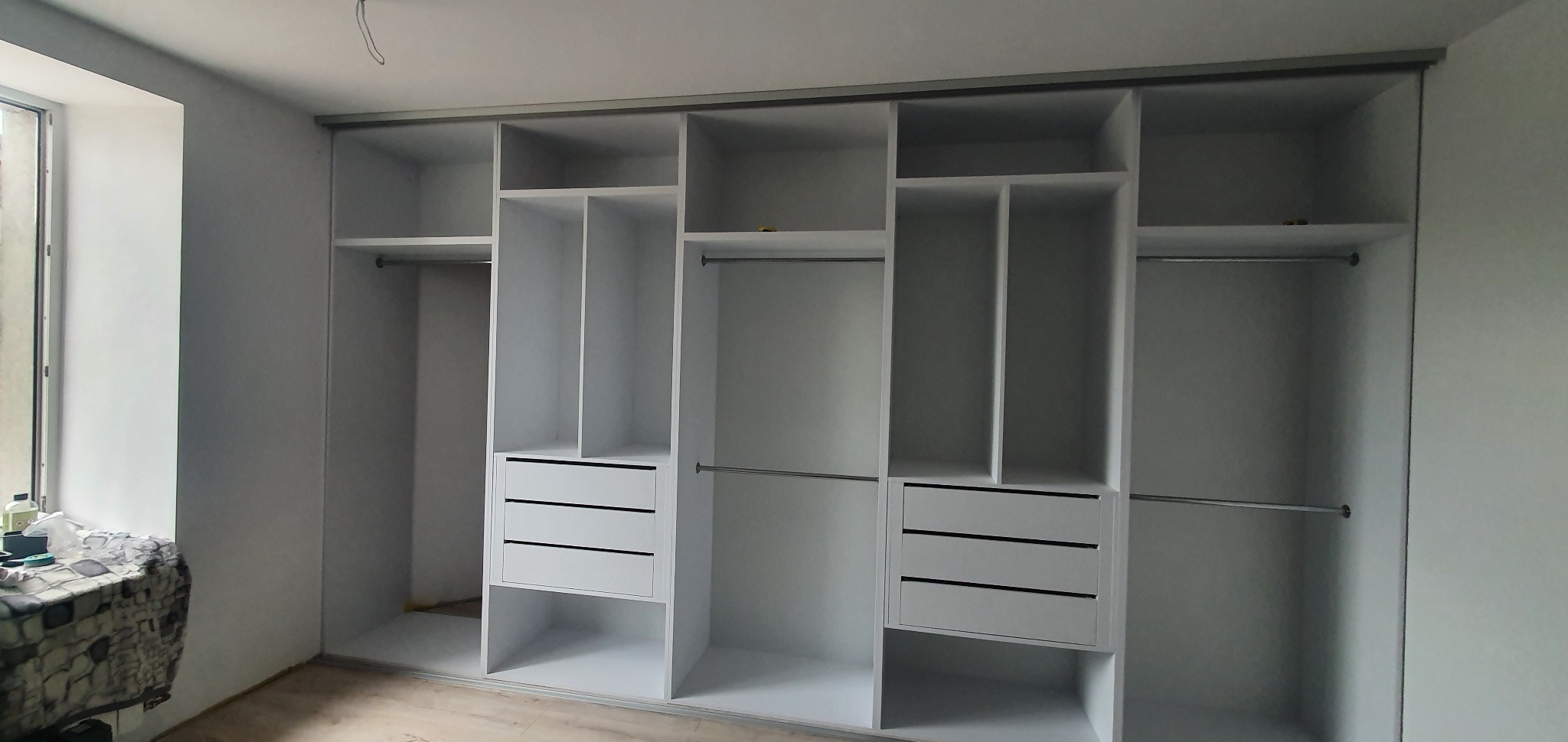 Wnętrze szafy przesuwnej z białymi frontami, widoczne półki, drążki na ubrania i trzy szuflady w jednej z sekcji, surowy stan wykończenia wnętrza.