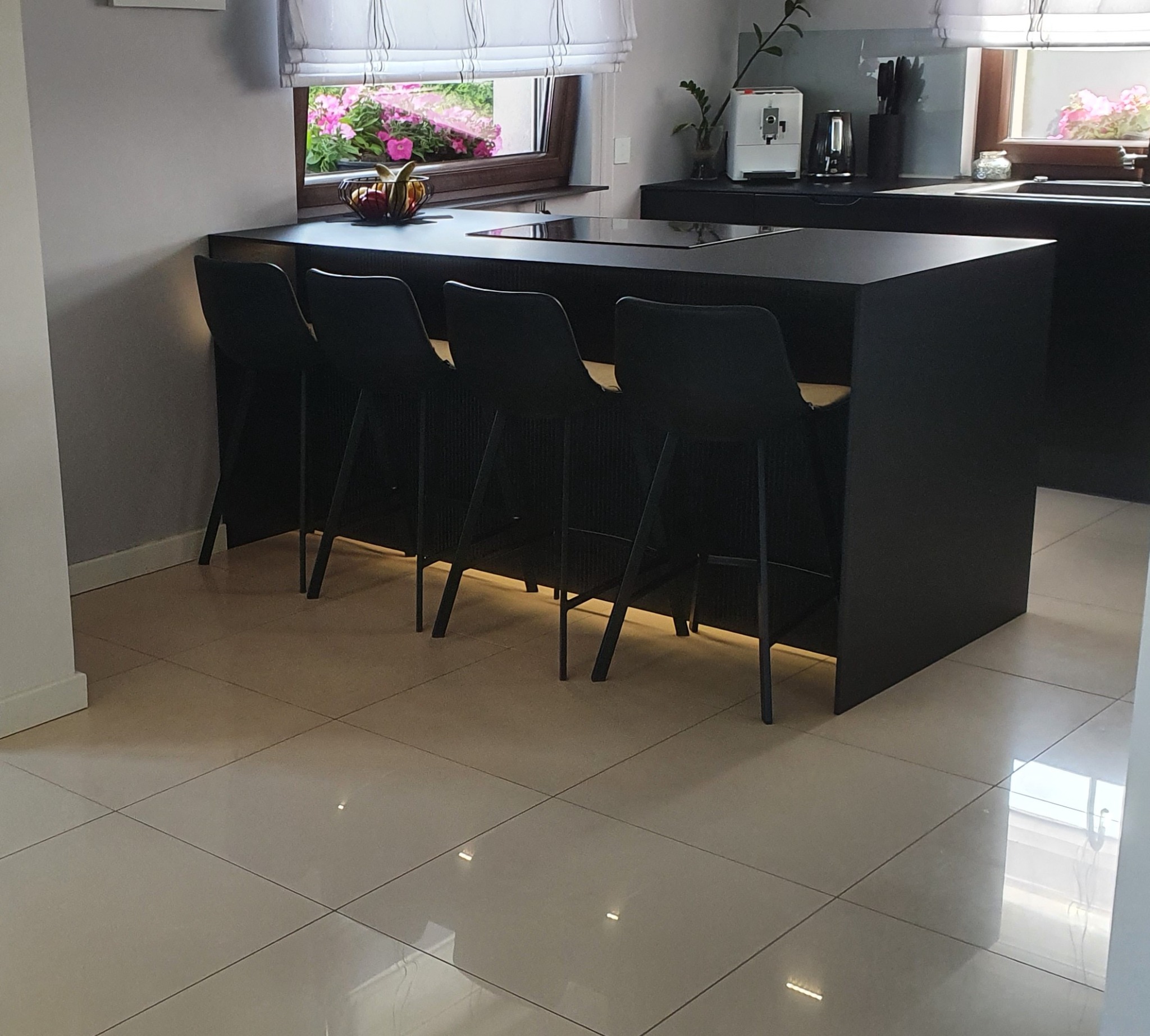 Nowoczesna, czarna wyspa kuchenna z płytą grzewczą, czterema hokerami z plecionym siedziskiem i oświetleniem LED u podstawy, widok na okno z kwiatami i ekspres do kawy w tle.