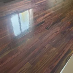 Świeżo wycyklinowana, błyszcząca podłoga z ciemnego drewna egzotycznego, z widocznym odbiciem okna i fragmentem białej listwy przypodłogowej.