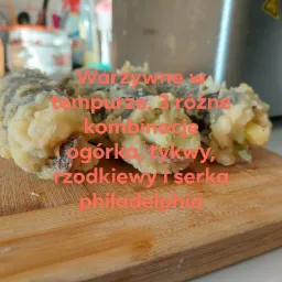 Trzy rolki sushi w tempurze z warzywami (ogórek, tykwa, rzodkiew) i serkiem Philadelphia, ułożone na drewnianej desce do krojenia, na tle srebrnego urządzenia.