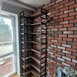 Kątowa półka z metalową ramą i drewnianymi półkami, wpasowana w narożnik pokoju z ceglaną ścianą i oknem balkonowym.