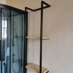 Metalowa konstrukcja z drewnianymi półkami zawieszona obok kabiny prysznicowej z czarnymi profilami.