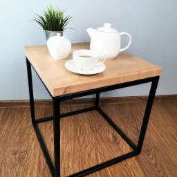 Stolik kawowy z blatem z jasnego drewna i czarną, metalową ramą, udekorowany białym serwisem do herbaty i doniczką z zieloną rośliną.