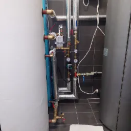 Dwa zbiorniki na wodę (biały i szary) z licznymi rurami, zaworami i manometrami, instalacja w kotłowni z szarą, kwadratową glazurą na ścianach i podłodze, widoczny fragment białego bloczka...