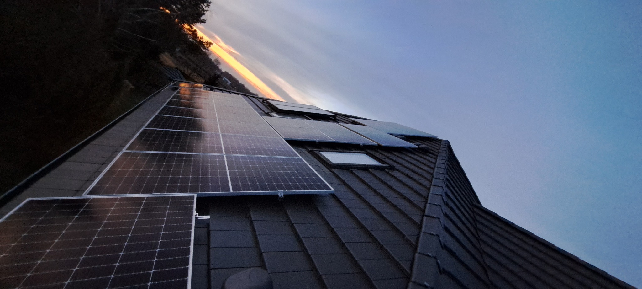 Panele słoneczne zamontowane na dachu domu z oknami dachowymi, widoczne na tle zachodzącego słońca.