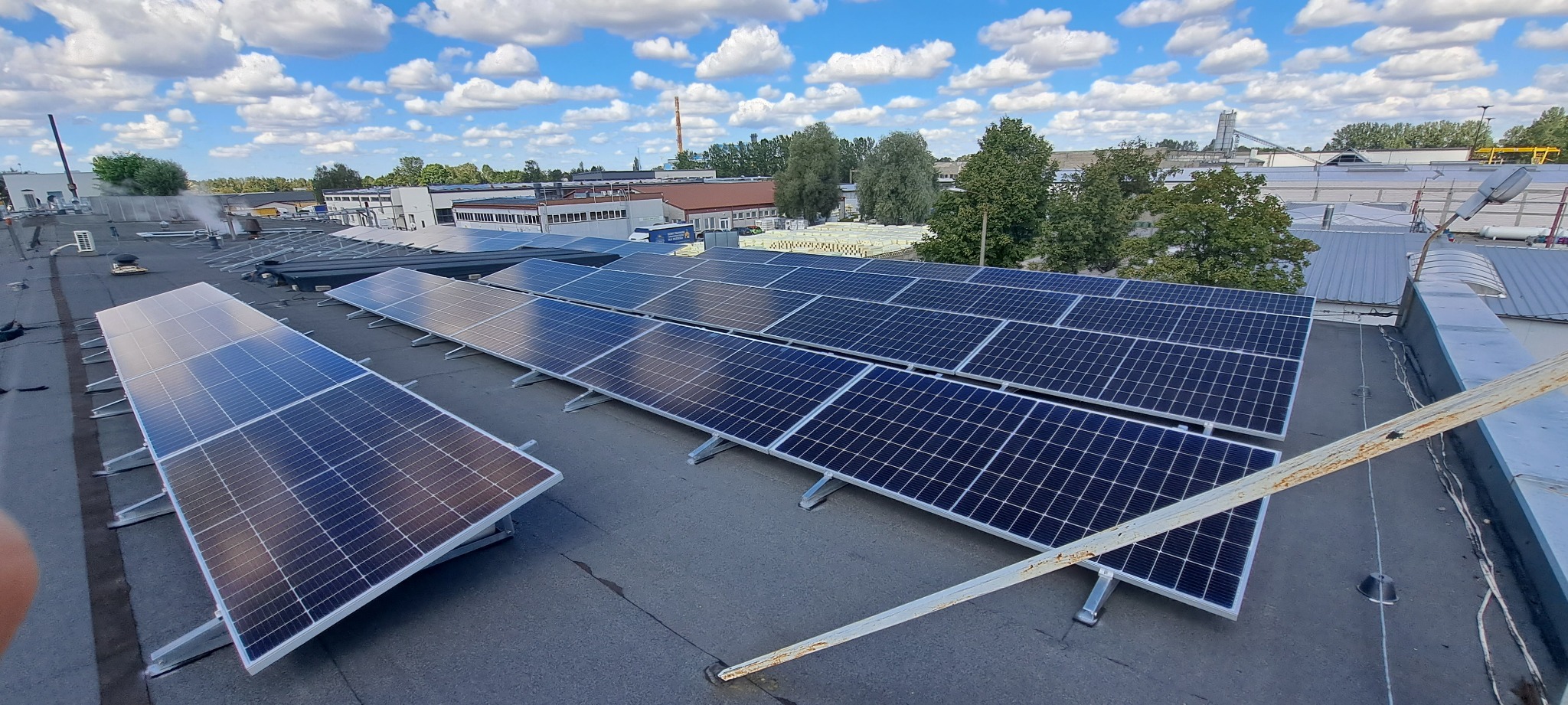 Instalacja paneli słonecznych na dachu budynku przemysłowego w Łodzi, widoczne niebo z chmurami i elementy konstrukcyjne dachu.