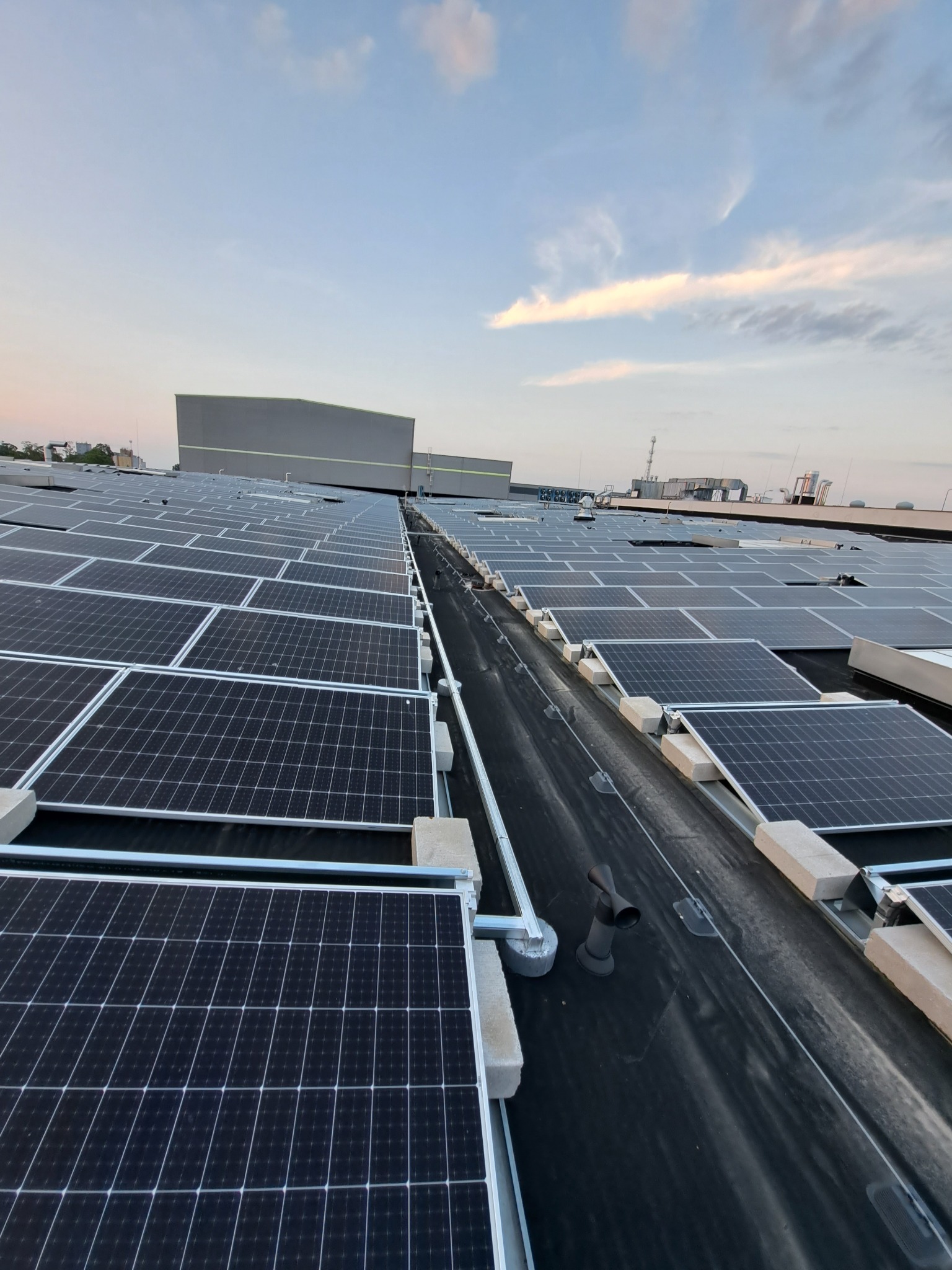 Rząd paneli fotowoltaicznych na płaskim dachu budynku przemysłowego, widoczne betonowe wsporniki i elementy wentylacji, w tle lekko zachmurzone niebo o zmierzchu.