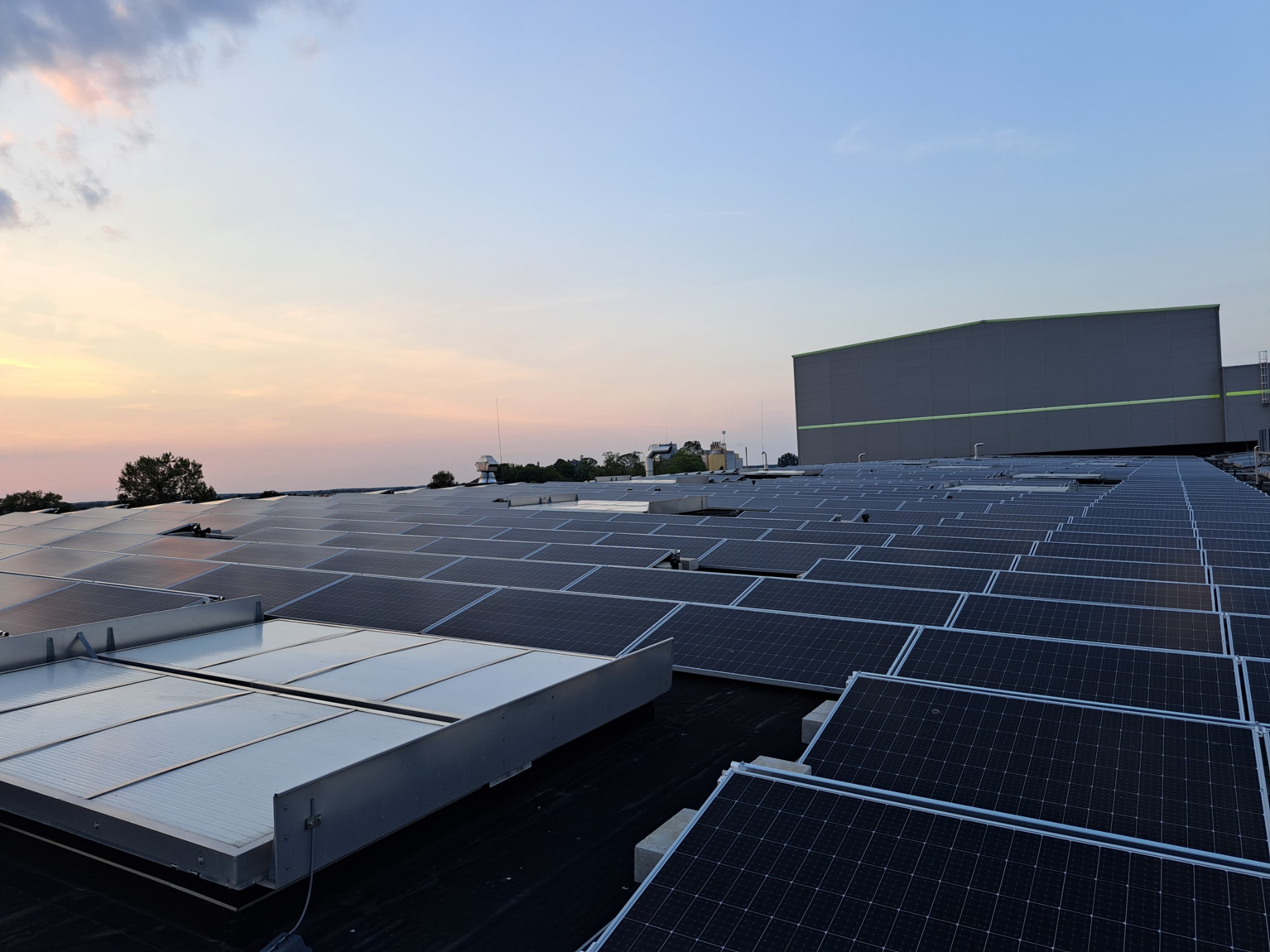 Rozległa instalacja paneli słonecznych na dachu budynku przemysłowego, widoczna konstrukcja wsporcza, zachodzące słońce barwi niebo na różowo.