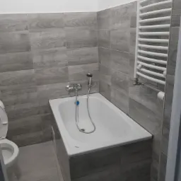 Wykończona łazienka z wanną obudowaną płytkami imitującymi drewno, białą toaletą i grzejnikiem drabinkowym, płytki ścienne w odcieniach szarości z subtelnym wzorem słojów drewna.