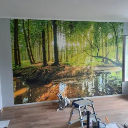 Fototapeta z motywem leśnym na ścianie, drabina malarska i akcesoria do malowania na podłodze z paneli.