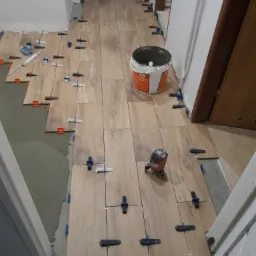 Układanie płytek imitujących drewno na podłodze w korytarzu, widoczne poziomice, wiadro z klejem i krzyżyki dystansowe.