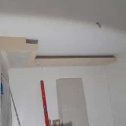 Wnętrze w trakcie prac wykończeniowych: częściowo zamontowany sufit podwieszany z płyt gipsowo-kartonowych, widoczne profile i poziomica.