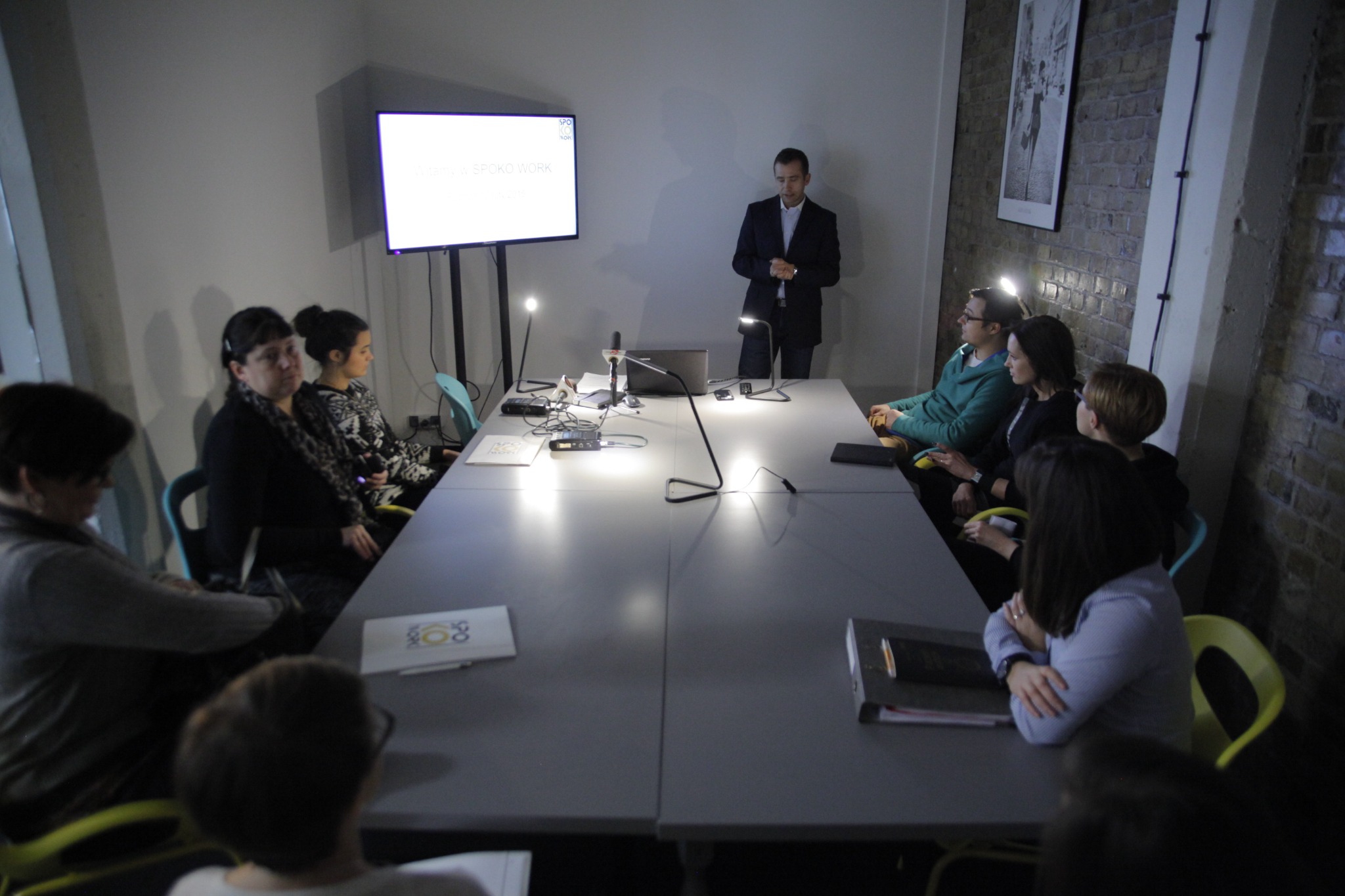 Sala konferencyjna w Poznaniu podczas prezentacji. Mężczyzna w garniturze stoi przed ekranem z napisem 'Crowd Work', otoczony przez grupę słuchaczy siedzących przy długim stole.