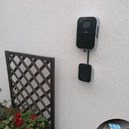 Na elewacji budynku zamontowana stacja ładowania pojazdów elektrycznych marki Green, obok drewniana kratka z czerwonymi kwiatami w donicy.