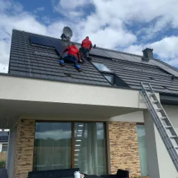 Dwóch monterów w czerwonych bluzach instaluje panele słoneczne na ciemnym dachu domu jednorodzinnego z oknami i elewacją z kamienia dekoracyjnego; drabina oparta o ścianę, błękitne niebo z chmurami...