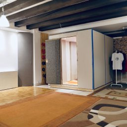 Studio Inżynierska - Wnętrze pomieszczenia z kabiną akustyczną z otwartymi drzwiami, wygłuszonymi panelami, widoczne mikrofony, artystyczny obraz na ścianie i ubrania na wieszaku.