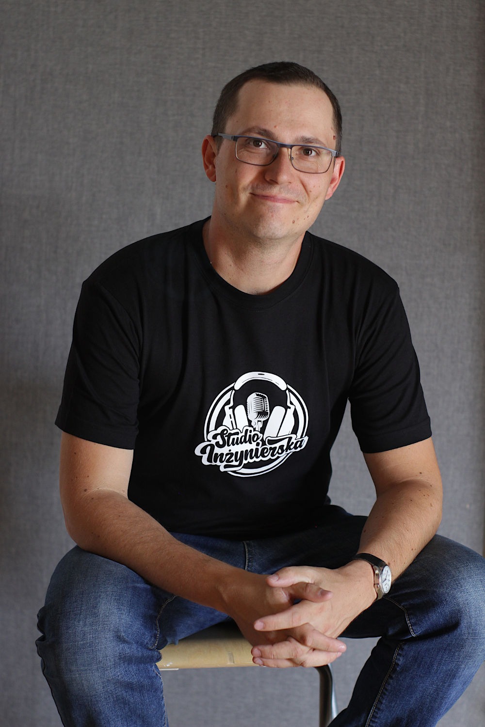 Mężczyzna w okularach i czarnej koszulce z logo studia nagraniowego, siedzący na krześle na szarym tle.