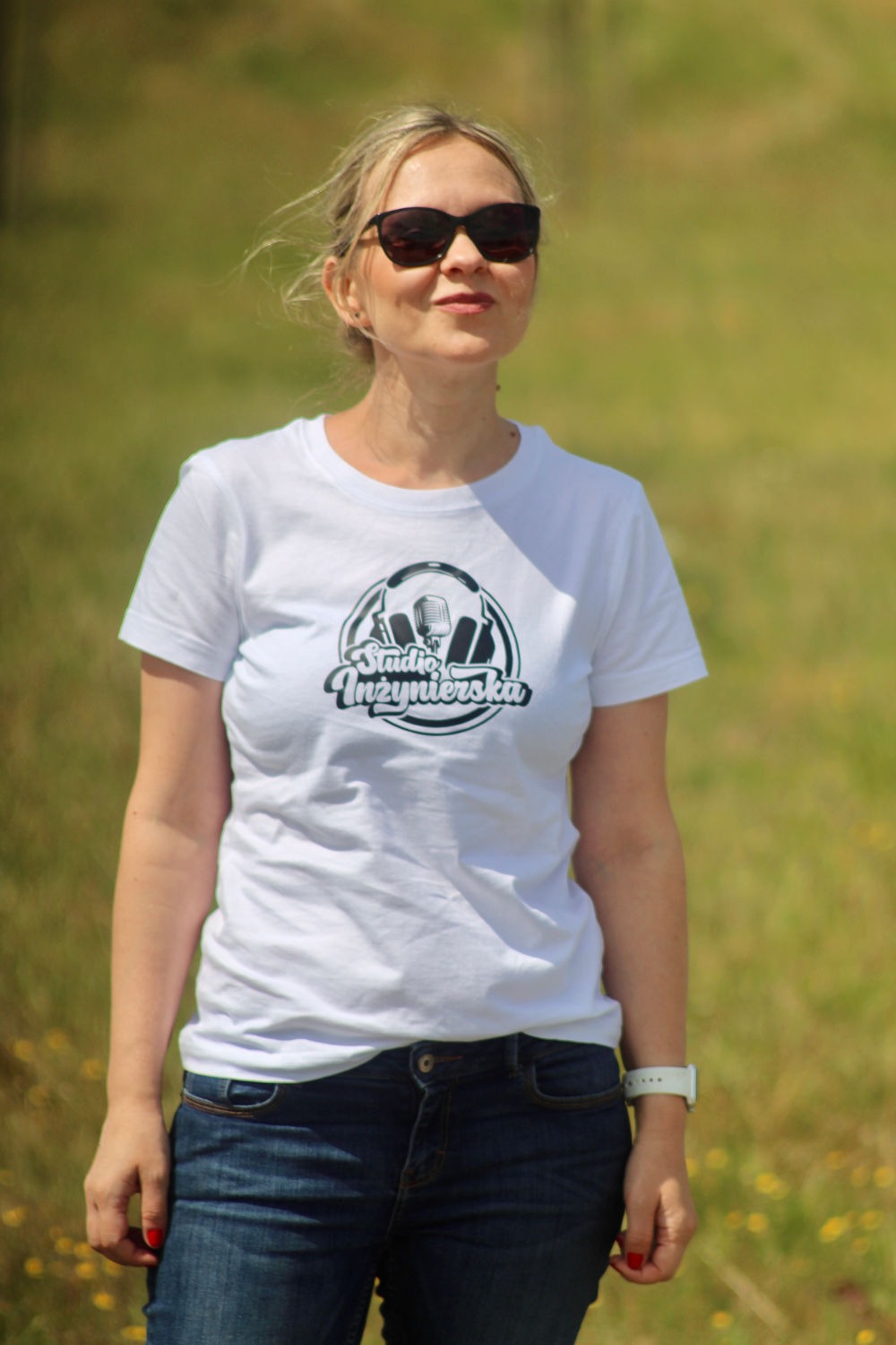 Kobieta w okularach przeciwsłonecznych i białej koszulce z logo firmy nagraniowej na tle zielonej trawy.