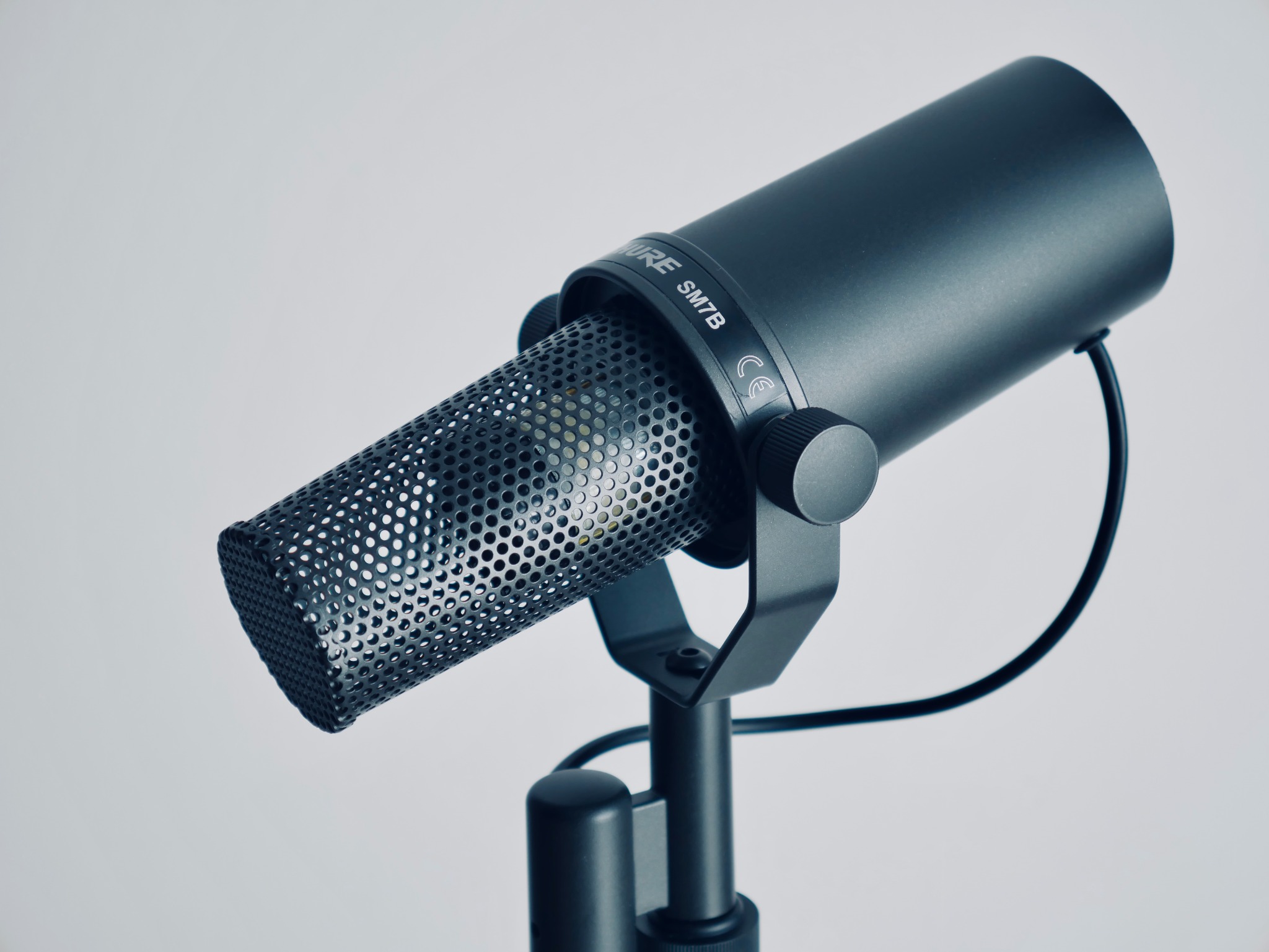 Czarny mikrofon Shure SM7B na statywie, widoczne logo i perforowana osłona, na jasnym tle.