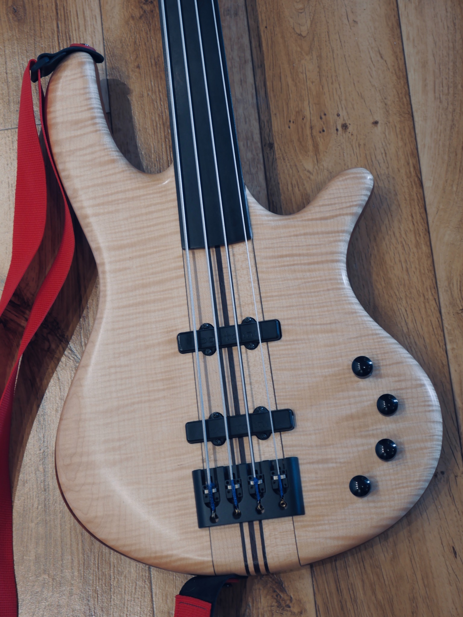 Fretless gitara basowa z jasnego drewna z czterema strunami i czerwonym paskiem, leżąca na drewnianej podłodze.