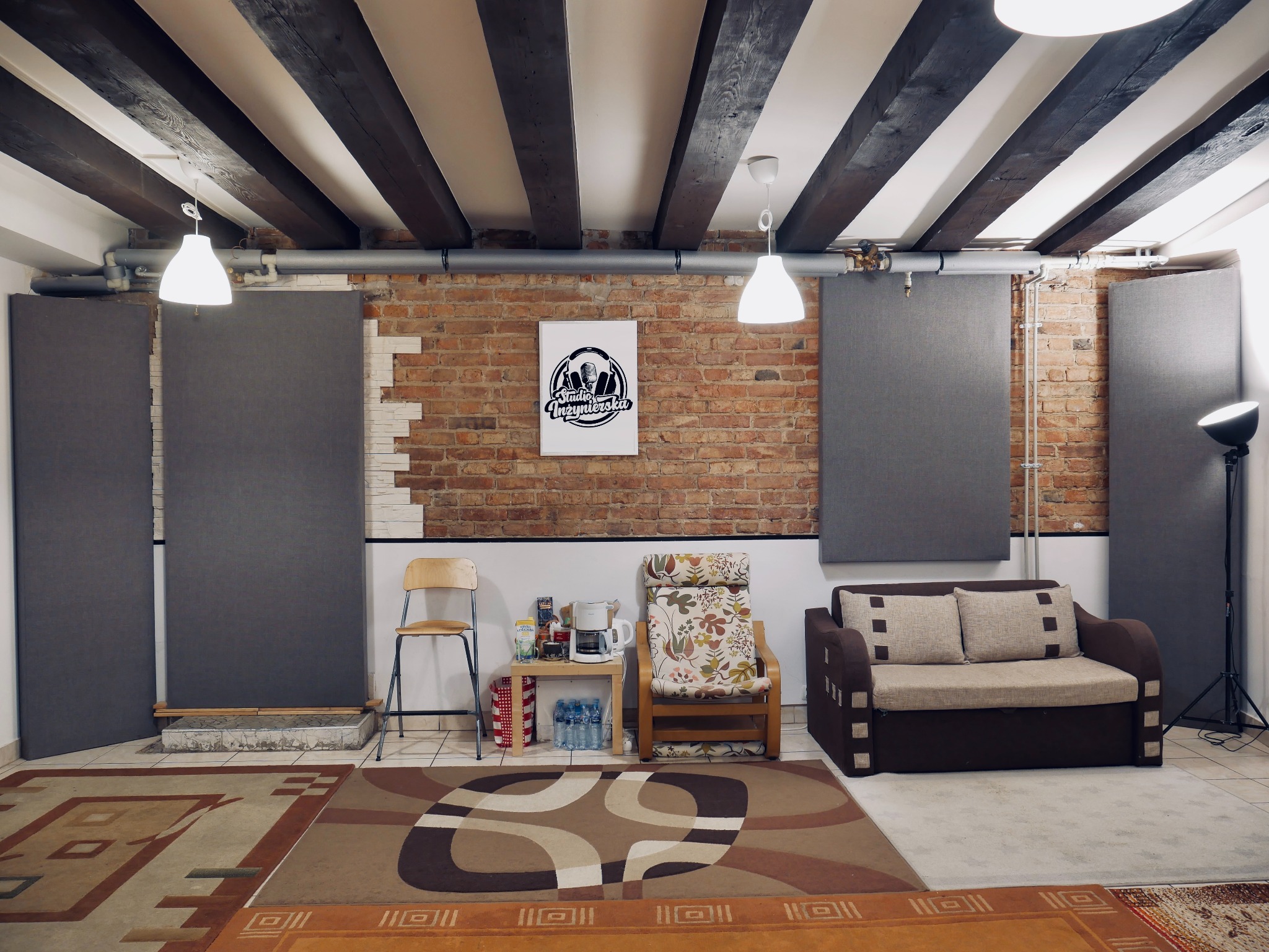 Wnętrze pokoju z panelami akustycznymi, ceglaną ścianą z plakatem z logo, sofą, fotelami i lampami sufitowymi