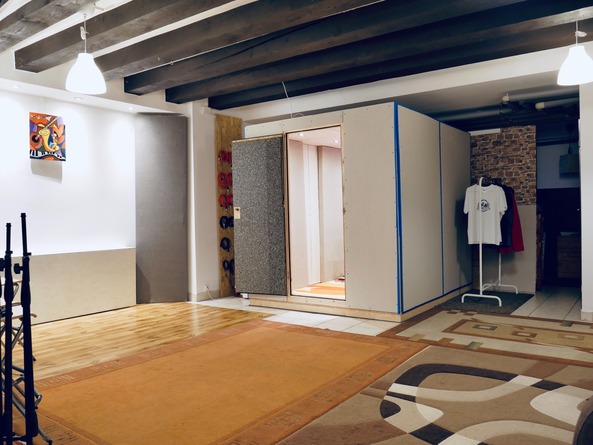 Wnętrze pomieszczenia z kabiną akustyczną z otwartymi drzwiami, wygłuszonymi panelami, widoczne mikrofony, artystyczny obraz na ścianie i ubrania na wieszaku.