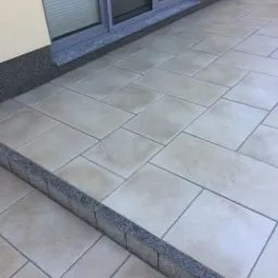 Jasne, prostokątne płyty tarasowe ułożone na zewnątrz budynku, z widocznym oknem i schodkiem wykończonym ciemnym granitem.