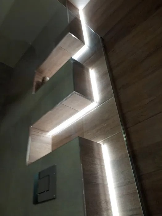 Półki wnękowe w łazience wyłożone płytkami imitującymi drewno, podświetlone paskami LED, z widocznym przyciskiem spłuczki w dolnej części kadru.