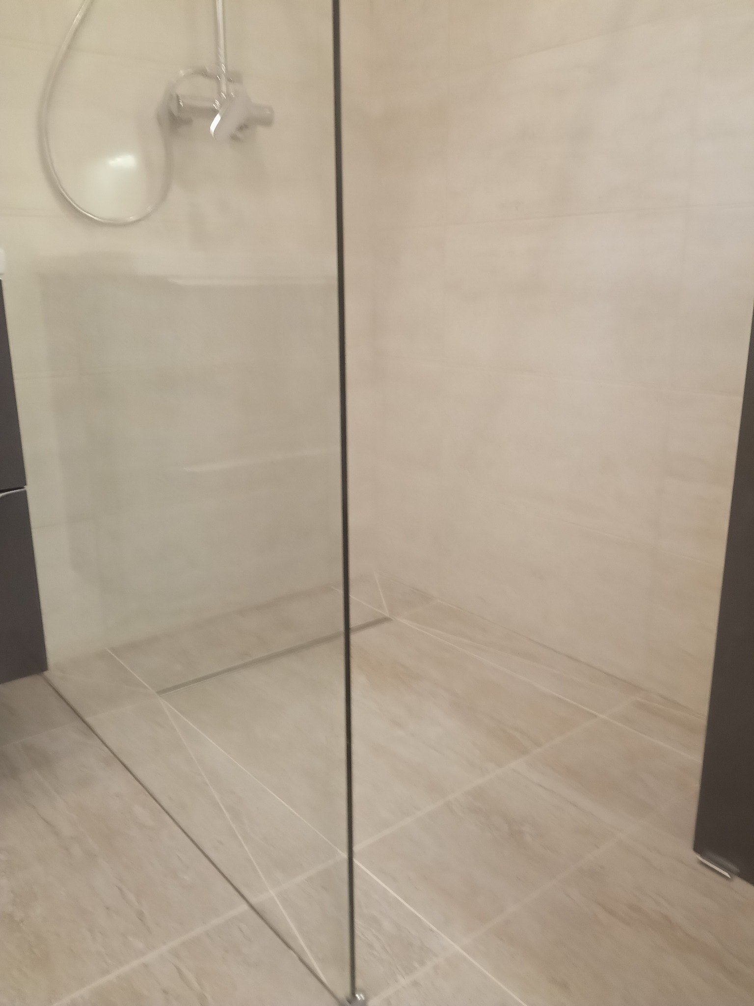 Szklana kabina prysznicowa z chromowaną armaturą na beżowych płytkach podłogowych i ściennych, widoczny fragment szafki pod umywalkę.