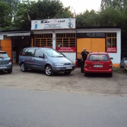 Warsztat samochodowy z zaparkowanymi samochodami osobowymi przed budynkiem; mężczyzna opiera się o czerwony samochód z tyłu, nad nim napis DIAGNOSTYKA KOMPUTEROWA.