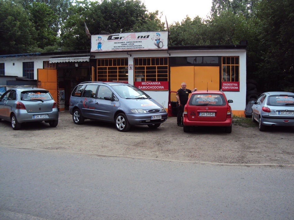 Warsztat samochodowy z zaparkowanymi samochodami osobowymi przed budynkiem; mężczyzna opiera się o czerwony samochód z tyłu, nad nim napis DIAGNOSTYKA KOMPUTEROWA.