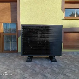 Czarna pompa ciepła Daikin Altherma 3 R ECH2O, zamontowana na zewnątrz domu z żółtą elewacją i brązowymi okiennicami, na tle kostki brukowej.