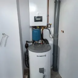 Biały piec De Dietrich z niebieskim zbiornikiem wyrównawczym, zamontowany na ścianie w surowym pomieszczeniu z szarą rurą kanalizacyjną. Częściowo jeszcze w folii ochronnej.