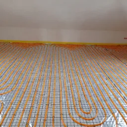 Pomarańczowe rury ogrzewania podłogowego ułożone spiralnie na siatce, gotowe do zalania wylewką. Żółta taśma przy ścianach. Widok z góry, ukośnie.