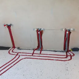 Wewnętrzne instalacje hydrauliczne w trakcie montażu, czerwone rury prowadzone w podłodze i wyprowadzone w ścianie, widoczne mocowania i pianka montażowa.