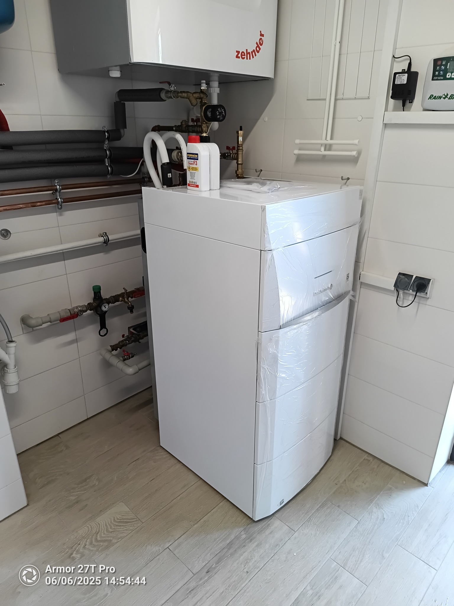 Nowy, biały piec grzewczy Zehnder, jeszcze w folii ochronnej, stoi w pomieszczeniu z instalacją hydrauliczną i białą glazurą. Widoczne rury i zawory.