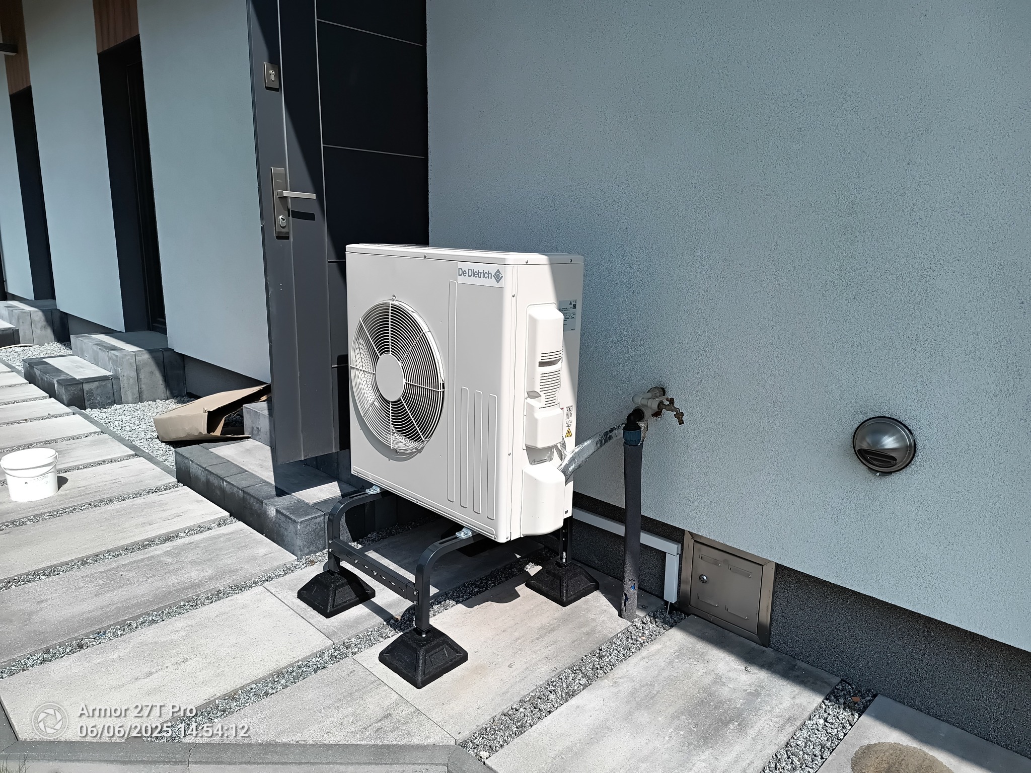 Biała pompa ciepła De Dietrich na czarnym stelażu, zainstalowana przy jasnej ścianie budynku. Widoczny kran i metalowa skrzynka rewizyjna.