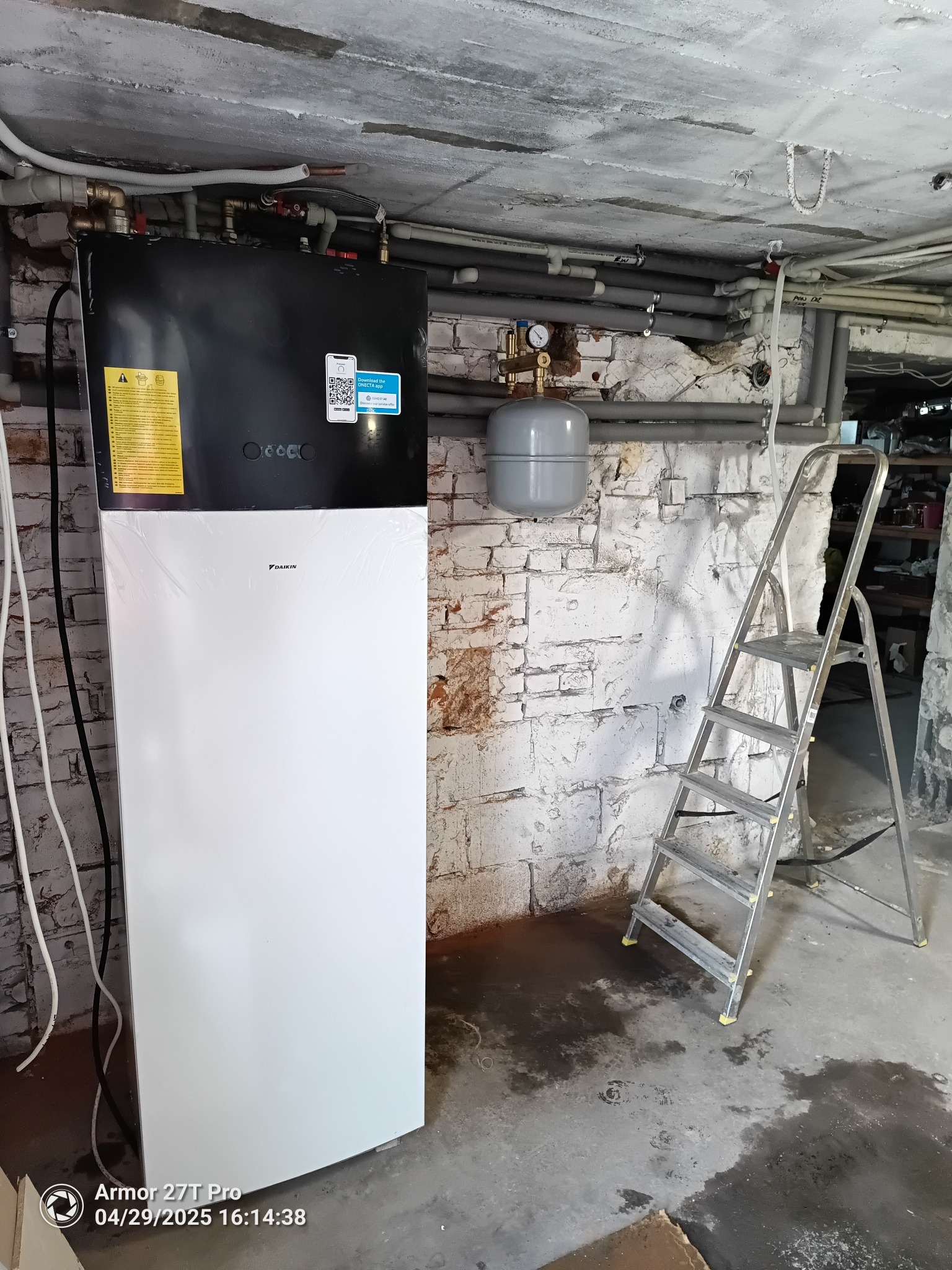 Biała pompa ciepła Daikin, z folią ochronną, zamontowana w piwnicy z instalacją hydrauliczną i drabiną. Widoczne rury i manometr.