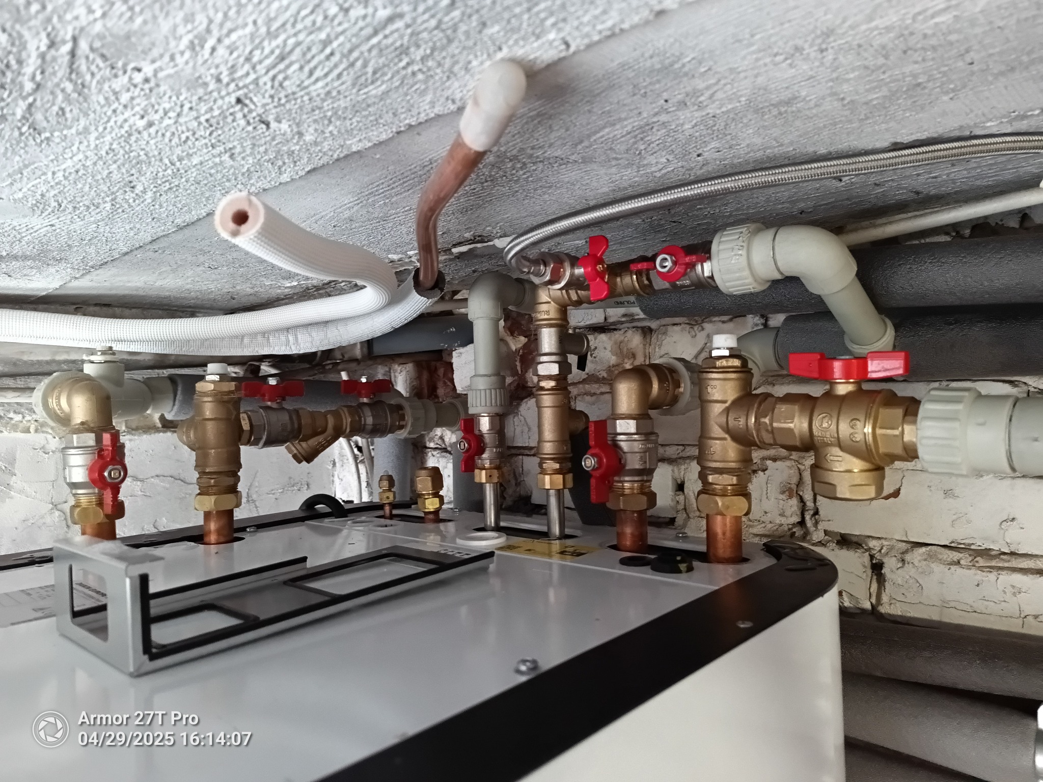 Zamknięte zawory z czerwonymi pokrętłami i rury miedziane, instalacja hydrauliczna pod sufitem w pomieszczeniu z ceglaną ścianą. Widok z dołu na biały bojler.