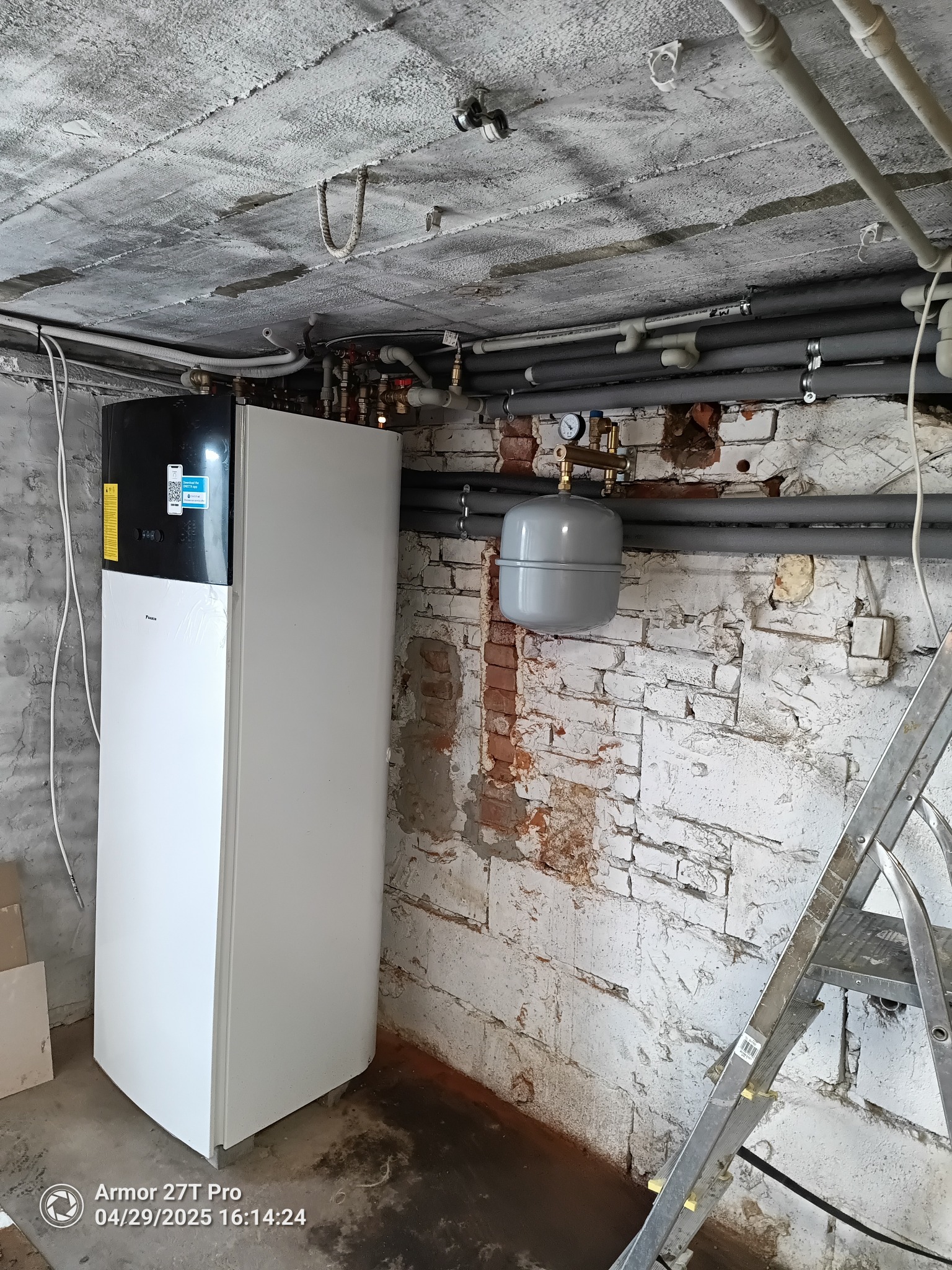 Biała pompa ciepła Daikin w piwnicy z rurami i zbiornikiem wyrównawczym na tle ceglanej ściany. Widoczna drabina i surowy betonowy strop.