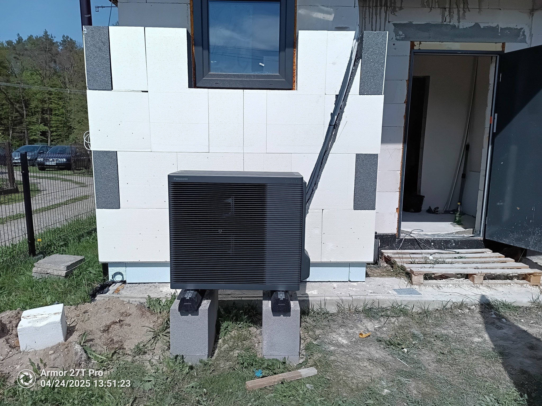 Czarna pompa ciepła Panasonic Aquarea zamontowana na betonowych blokach przed budynkiem w trakcie budowy. Widoczne okno i niedokończone ściany z białych bloków.