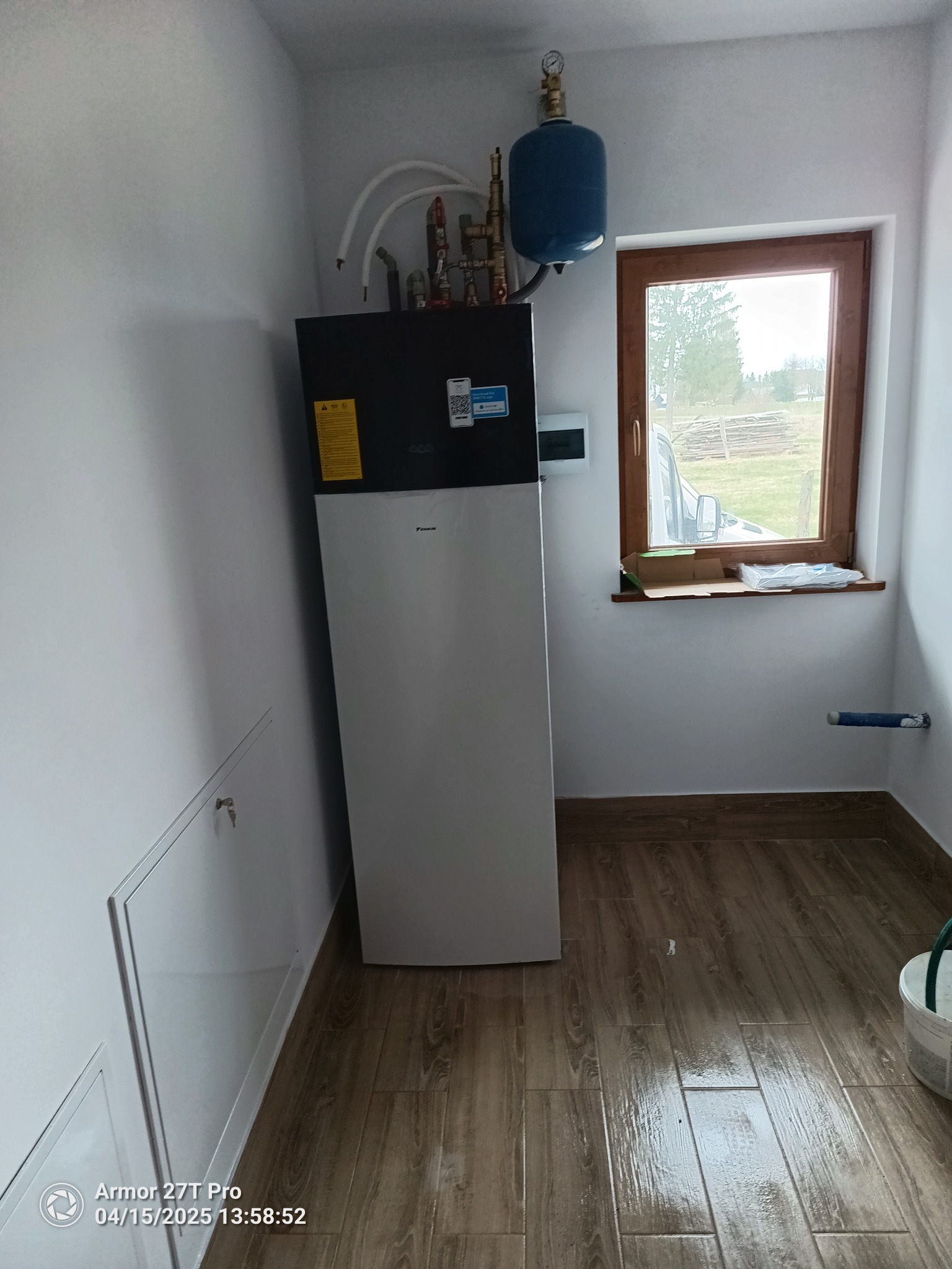 Nowoczesna pompa ciepła Daikin zamontowana w pomieszczeniu z drewnopodobną podłogą i oknem. Instalacja hydrauliczna z zbiornikiem wyrównawczym.