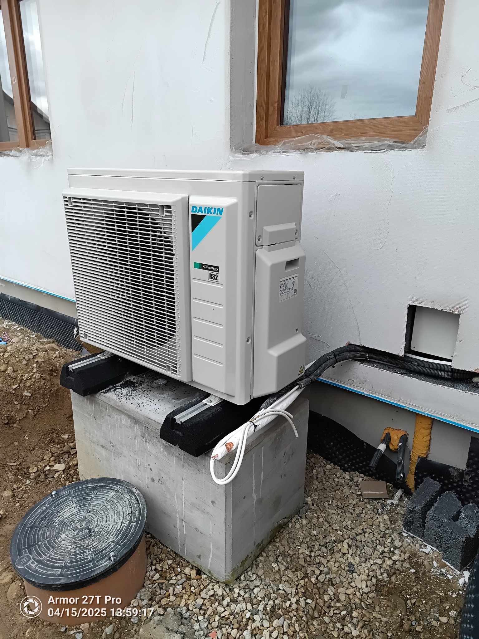 Zewnętrzna jednostka pompy ciepła Daikin zamontowana na betonowym fundamencie przy ścianie budynku z widocznymi przewodami i elementami instalacji.