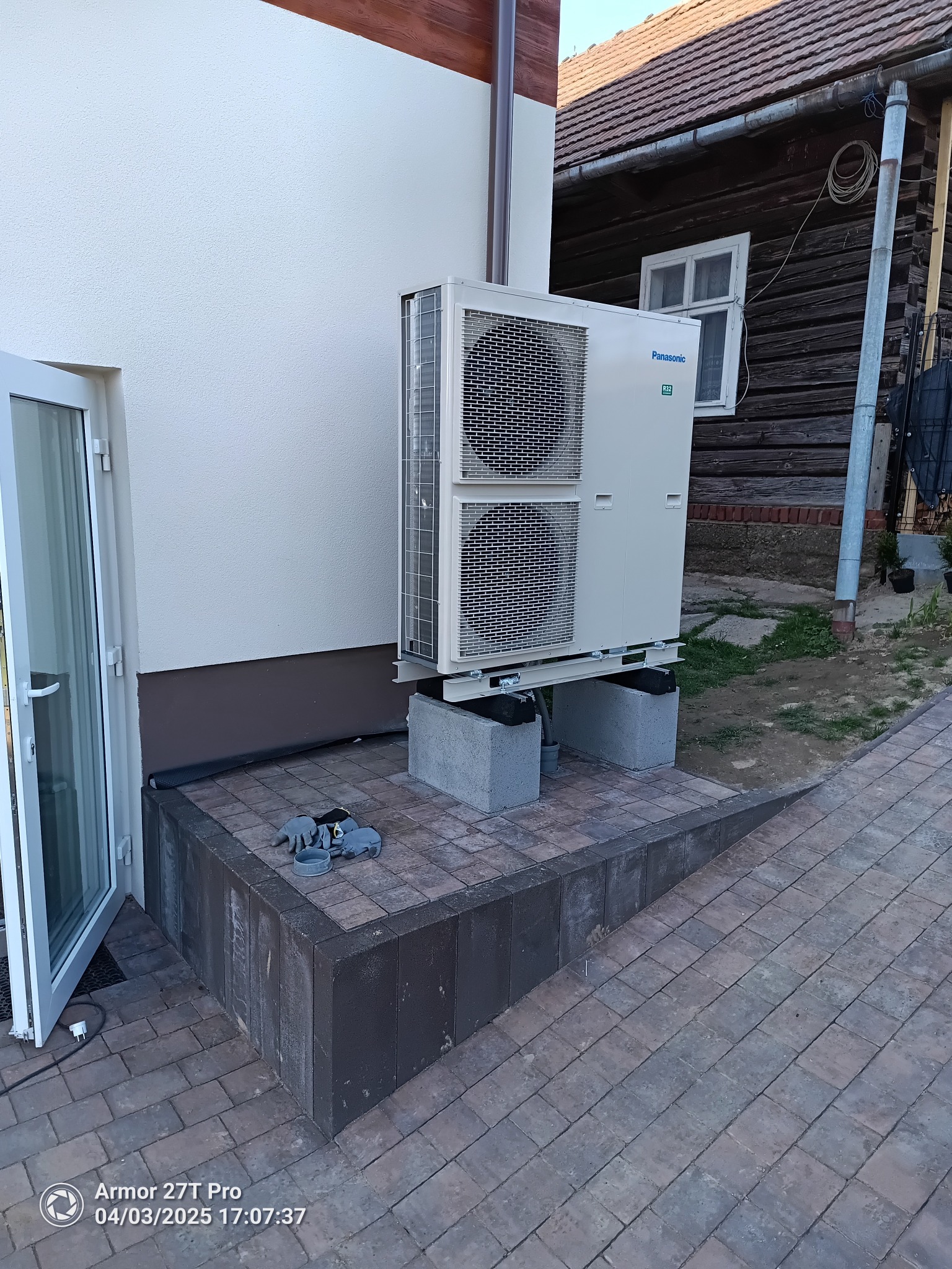 Nowoczesna pompa ciepła Panasonic zamontowana na betonowych blokach przy ścianie budynku z ceglaną kostką brukową i otwartymi drzwiami wejściowymi.
