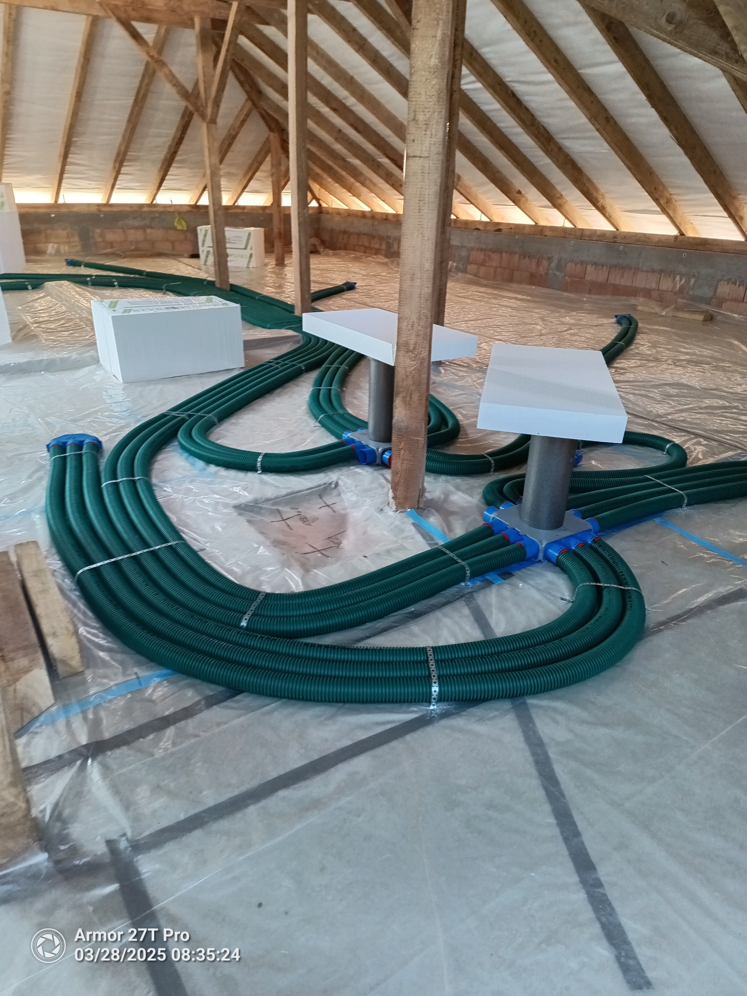 Wnętrze poddasza z elementami instalacji rekuperacji: zielone rury wentylacyjne podłączone do białych skrzynek rozdzielczych. Widoczne drewniane elementy konstrukcyjne dachu.