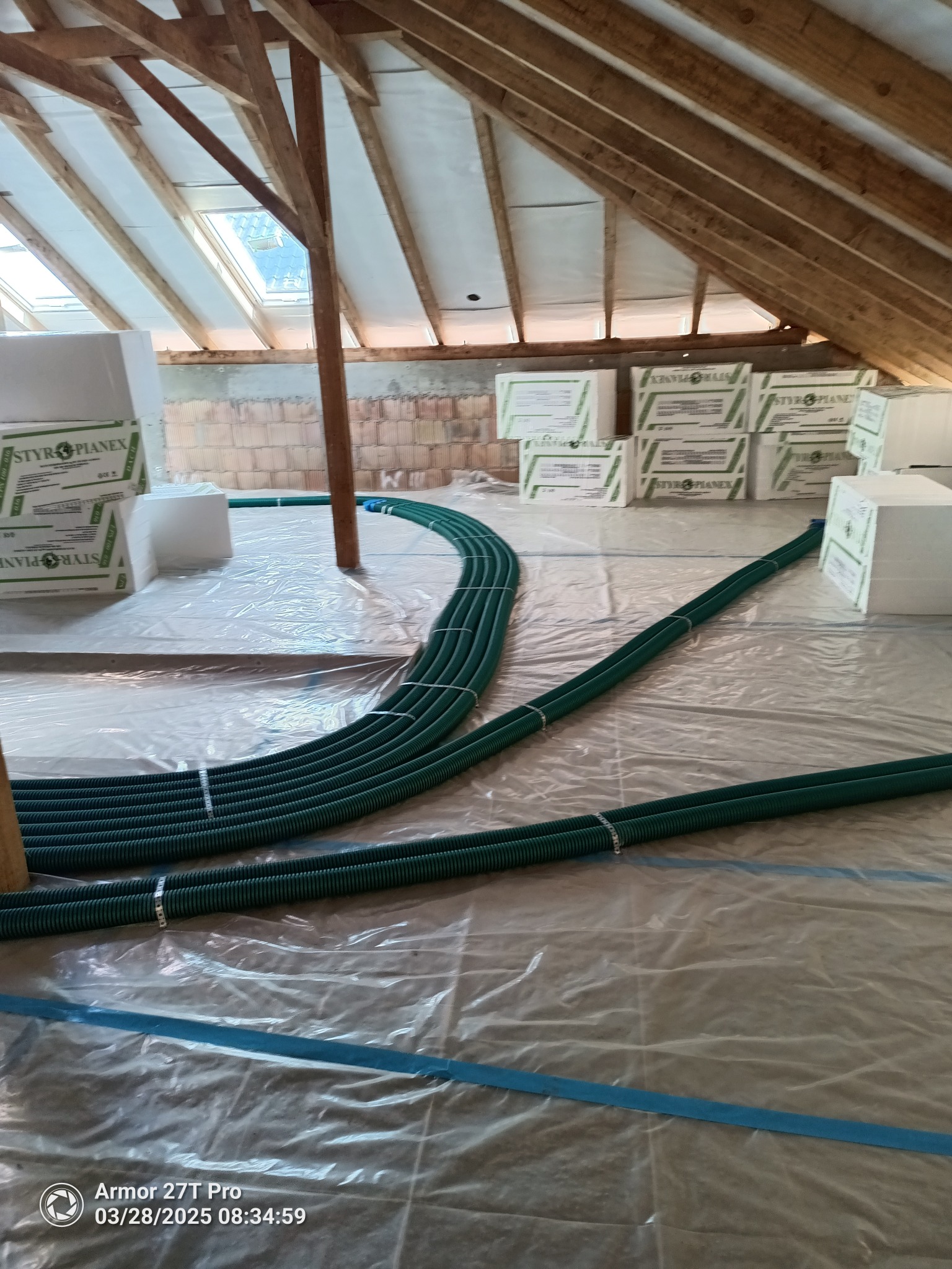 Zielone rury rekuperacji ułożone na poddaszu, widoczne pakiety styropianu i konstrukcja dachu z oknami. Instalacja w trakcie realizacji, zabezpieczona folią.