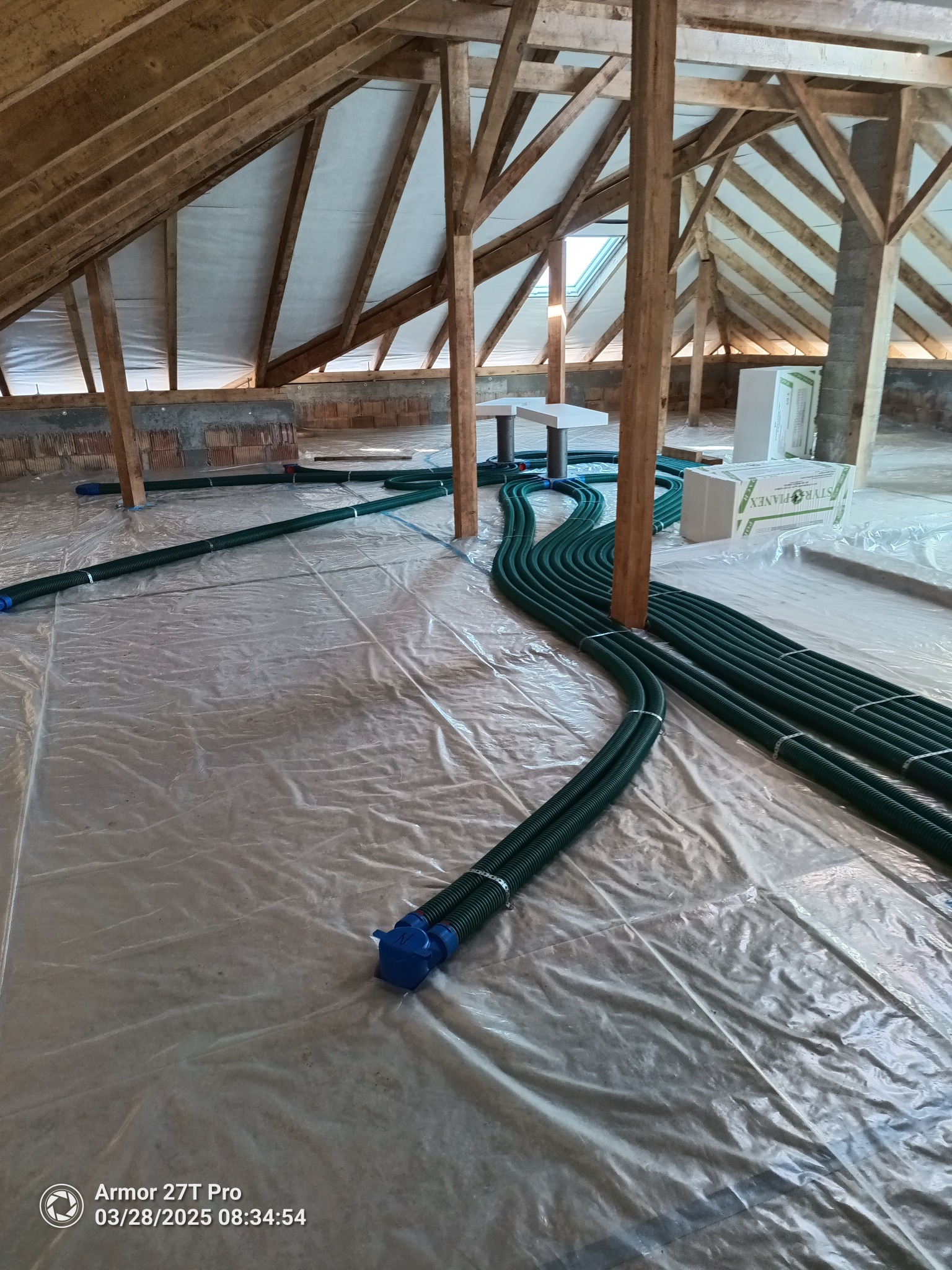 Zielone rury wentylacyjne ułożone na folii w surowym poddaszu z drewnianą konstrukcją. Widoczne elementy instalacji i materiały izolacyjne.